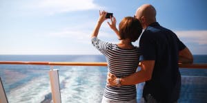 Middle aged couple on a cruise ship looking out to a blue ocean and sunshine. The lady holds a phone up, taking a picture of the view.